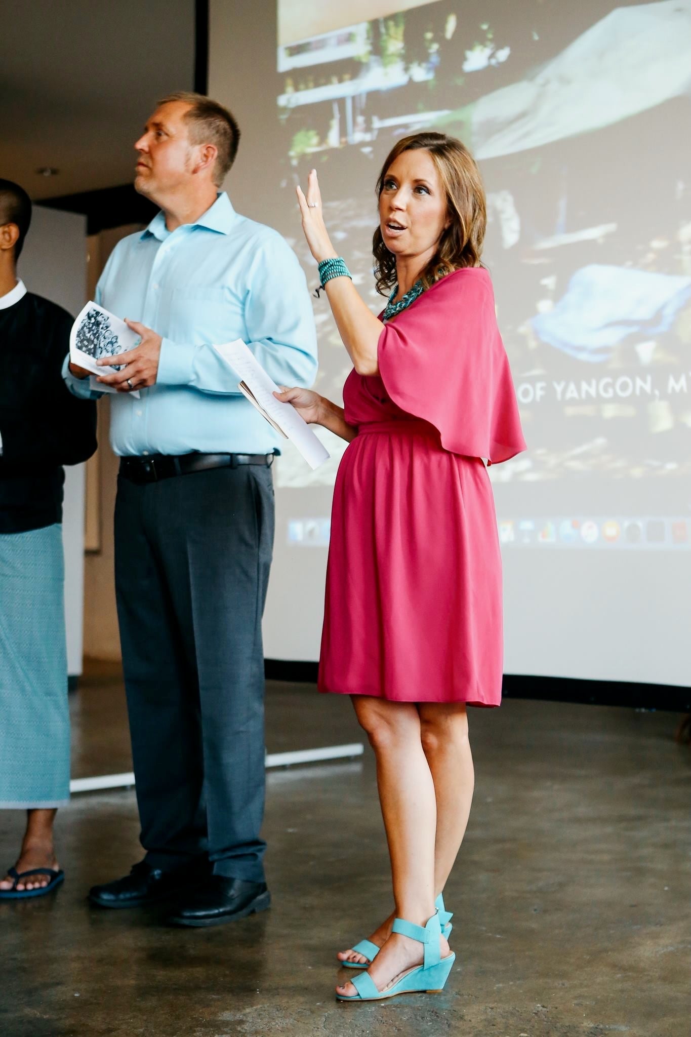 Woman in a pink dress speaking at an event with a man holding an award in front of a screen.